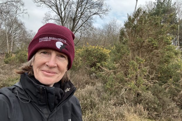 Selfie photo of Sarah in her burgundy "Thames Basin Heaths" woolly hat and heathland behind.