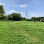 Photograph of a grassy path across a wide green meadow. A pylon stands prominently.