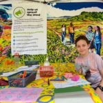 Photo of a yound girl doing origami at a table. Behind her is a colourful backdrop of a painting of people walking on a heath.