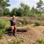 Photo of a man sitting on a fallen tree trunk. At one end is a large pile of pine needles...a Wood Ant nest.