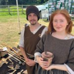 Photo of two young people dressed in Bronze Age style clothing, there are axes and implements on the ground.