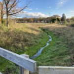 Photo of a small stream flowing through an open grass area with a wooden fence in front